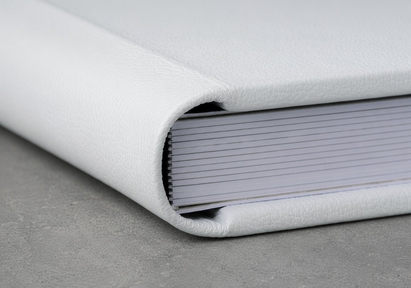 Buchrücken eines weißen Leder-Fotoalbums in Nahaufnahme, Layflat-Bindung und Lederstitching auf grauem Stein sichtbar.