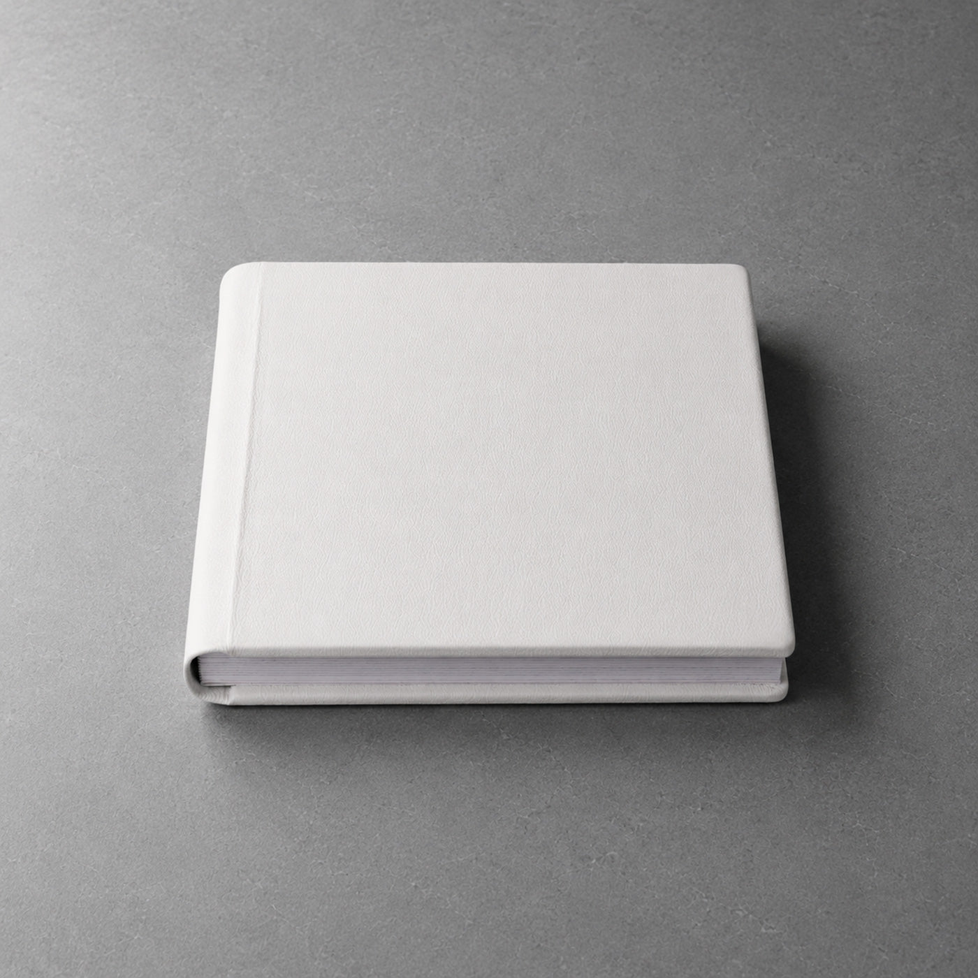 Geschlossenes Fotoalbum aus schneeweißem Vollnarbenleder, 30×30 cm, Frontansicht auf matter Steinplatte, weiches Seitenlicht von links.